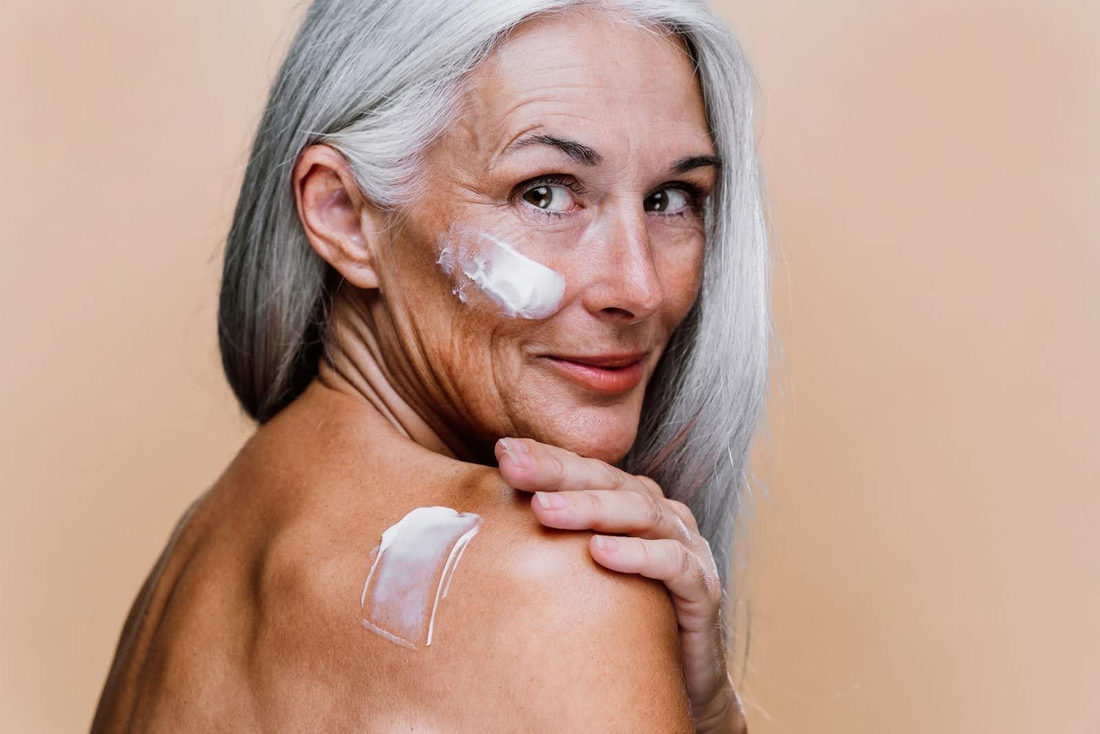 Older woman with white hair and clear skin applies lotion to face and shoulder